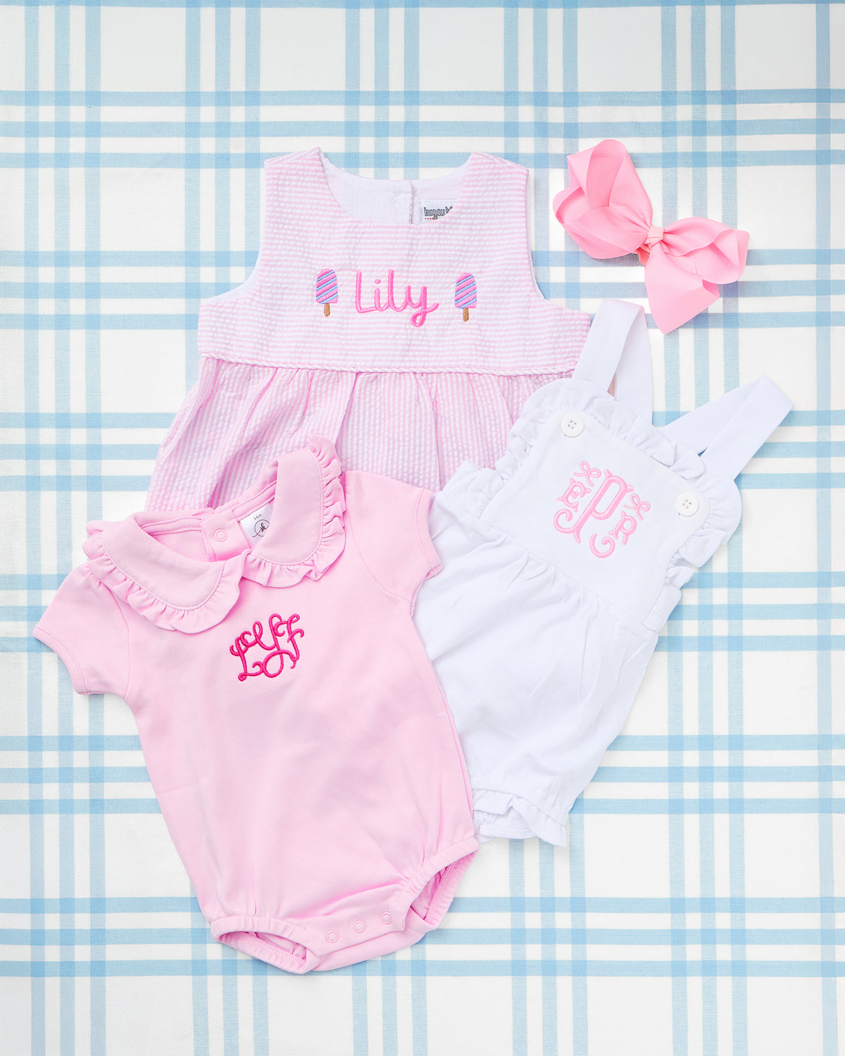 Flat lay of three adorable baby girl outfits on a soft blue plaid backdrop: a light pink Seersucker Bubble with Motif embroidered with “Lily” and sweet popsicle designs, a crisp white Cotton Sunsuit with elegant pink monogramming, and a pink Custom Name Short Sleeve Peter Pan Collar Bubble featuring a delicate ruffle collar and matching monogram. A coordinating pink bow adds the finishing touch to this charming and personalized summer-ready ensemble.