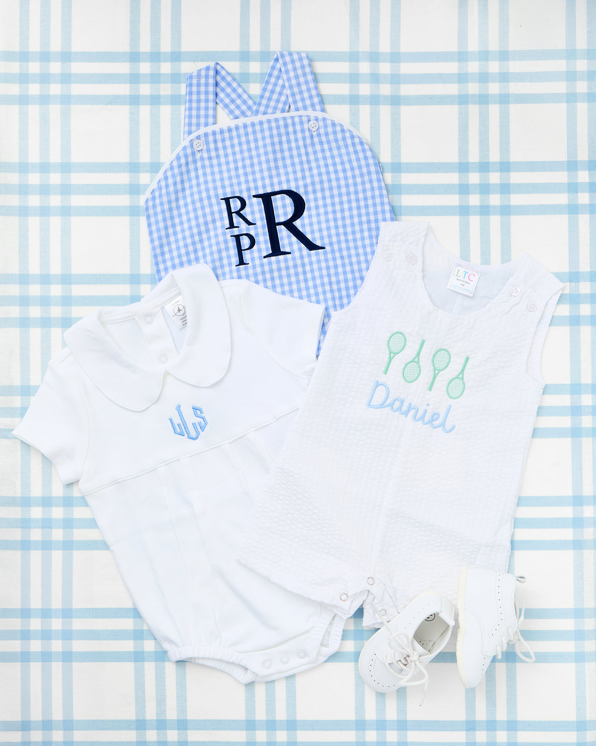 Flat lay of three charming baby outfits styled on a soft blue plaid background. Centered is a Custom Name Short Sleeve Peter Pan Collar Bubble in crisp white, personalized with the initials “WJS” embroidered in a soft blue thread on the collar, offering a classic and timeless look. Accompanying the bubble are a blue gingham Gingham Sunsuit with bold navy monogram “RPR” and a white Seersucker Jon Jon featuring green and blue tennis racket motifs with the name “Daniel.” Perfect for a polished, Southern-inspir