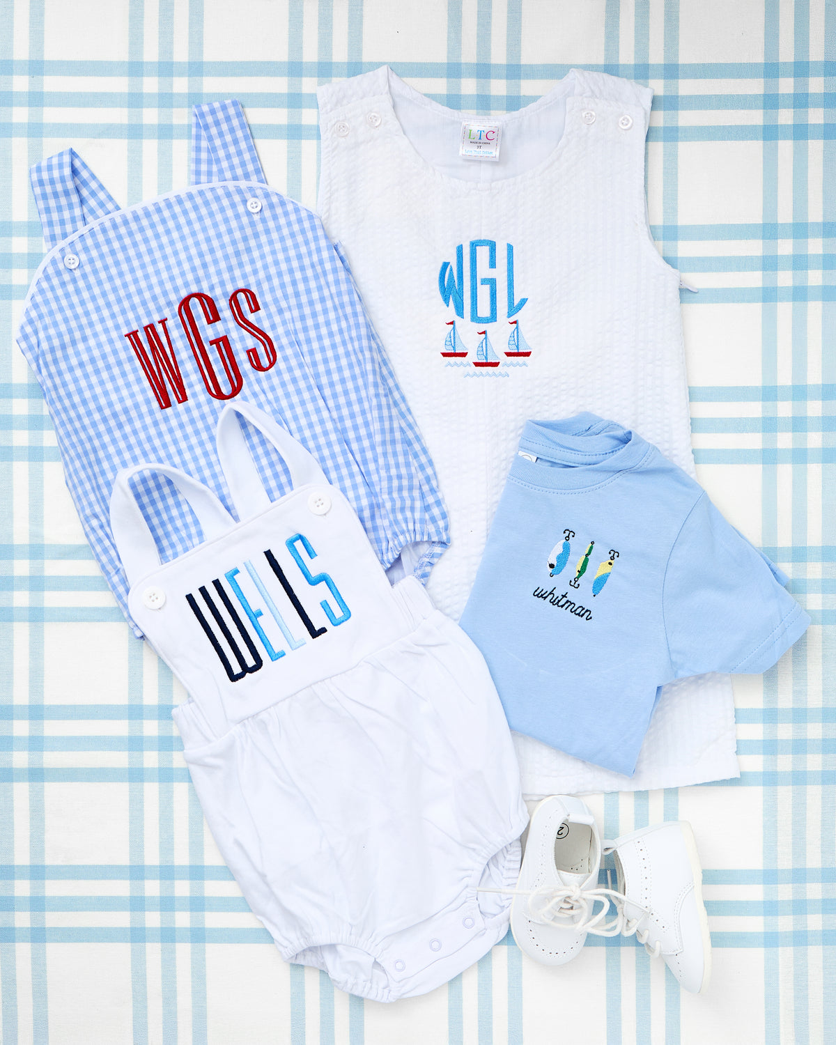 Flat lay featuring a collection of custom baby apparel from Rainey Laine, including a personalized blue Short Sleeve Striped Bubble embroidered with the name "whitman" and colorful fishing lures. Surrounding the bubble are a Gingham Sunsuit with red monogram "WGS," a white Cotton Sunsuit with bold "WELLS" lettering, and a Seersucker Jon Jon with a sailboat motif and initials "WGL." White baby shoes complete this preppy, personalized look perfect for summer outings.