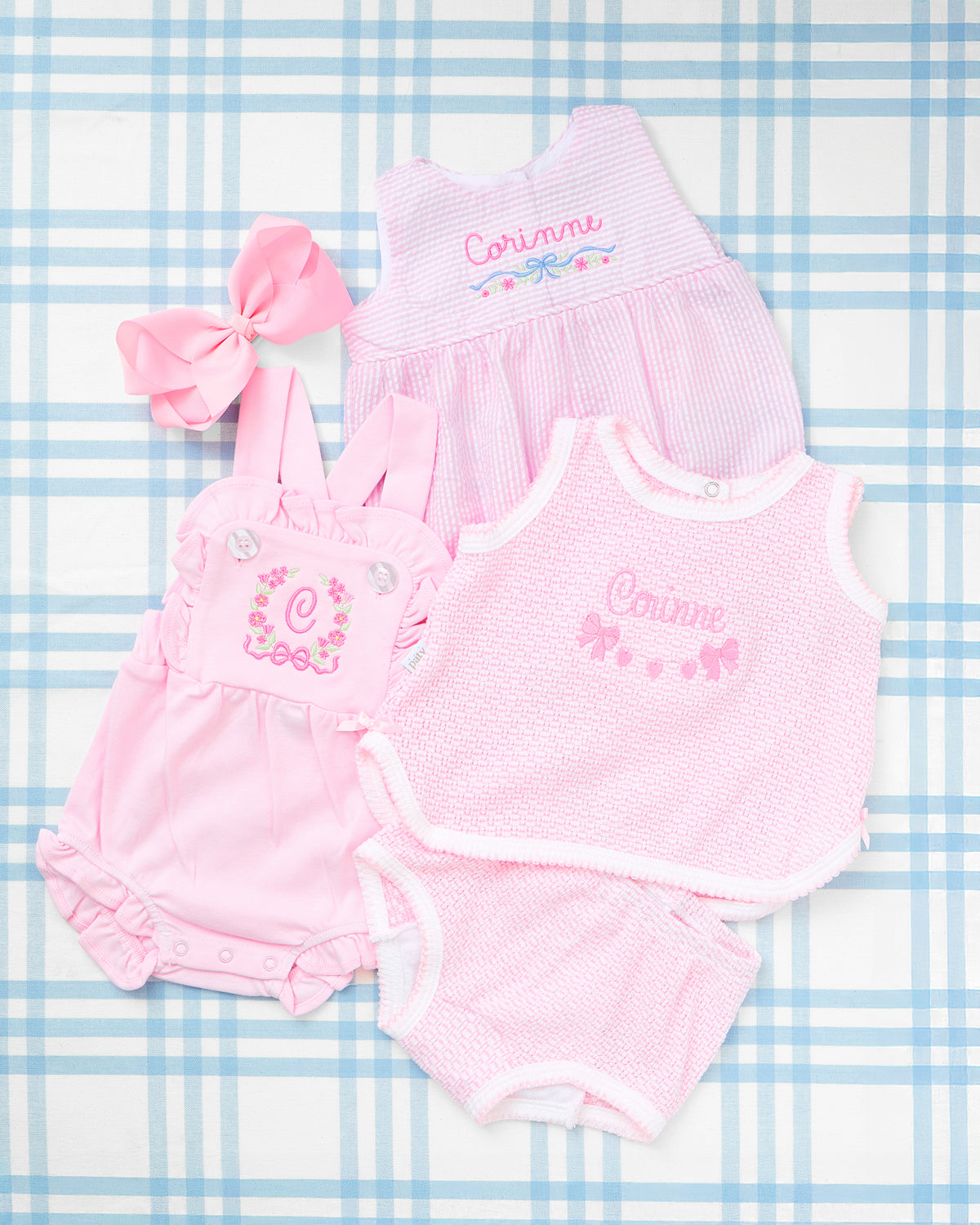 Flat lay of three charming baby girl outfits styled on a blue plaid background: a pink Seersucker Bubble with Motif embroidered with "Corinne" and delicate floral accents, a Cotton Sunsuit with Patriotic Motif featuring ruffle straps and a floral monogrammed “C”, and a cozy pink Cotton Sunsuit set with matching top and bloomers, personalized with “Corinne” and a playful bow design. A coordinating pink bow completes the sweet, preppy look—perfect for spring and summer occasions.