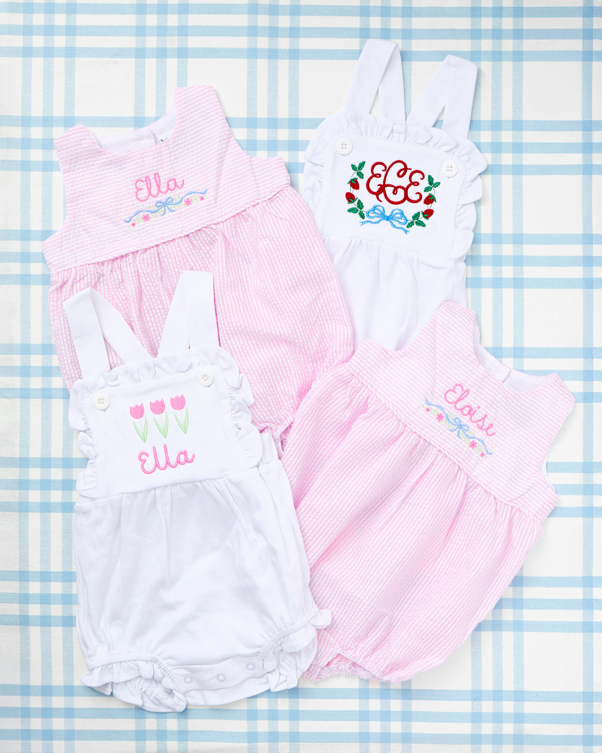 Four adorable baby outfits are displayed on a light blue plaid backdrop, including two Seersucker Bubbles with Motif in pink featuring custom embroidery with floral and bow accents and the names Ella and Eloise, along with two crisp white Cotton Sunsuits with Patriotic Motif—one with tulip embroidery and the name Ella, and the other with a striking red, white, and blue floral monogram. These classic pieces blend Southern charm with playful personalization, perfect for warm-weather celebrations.