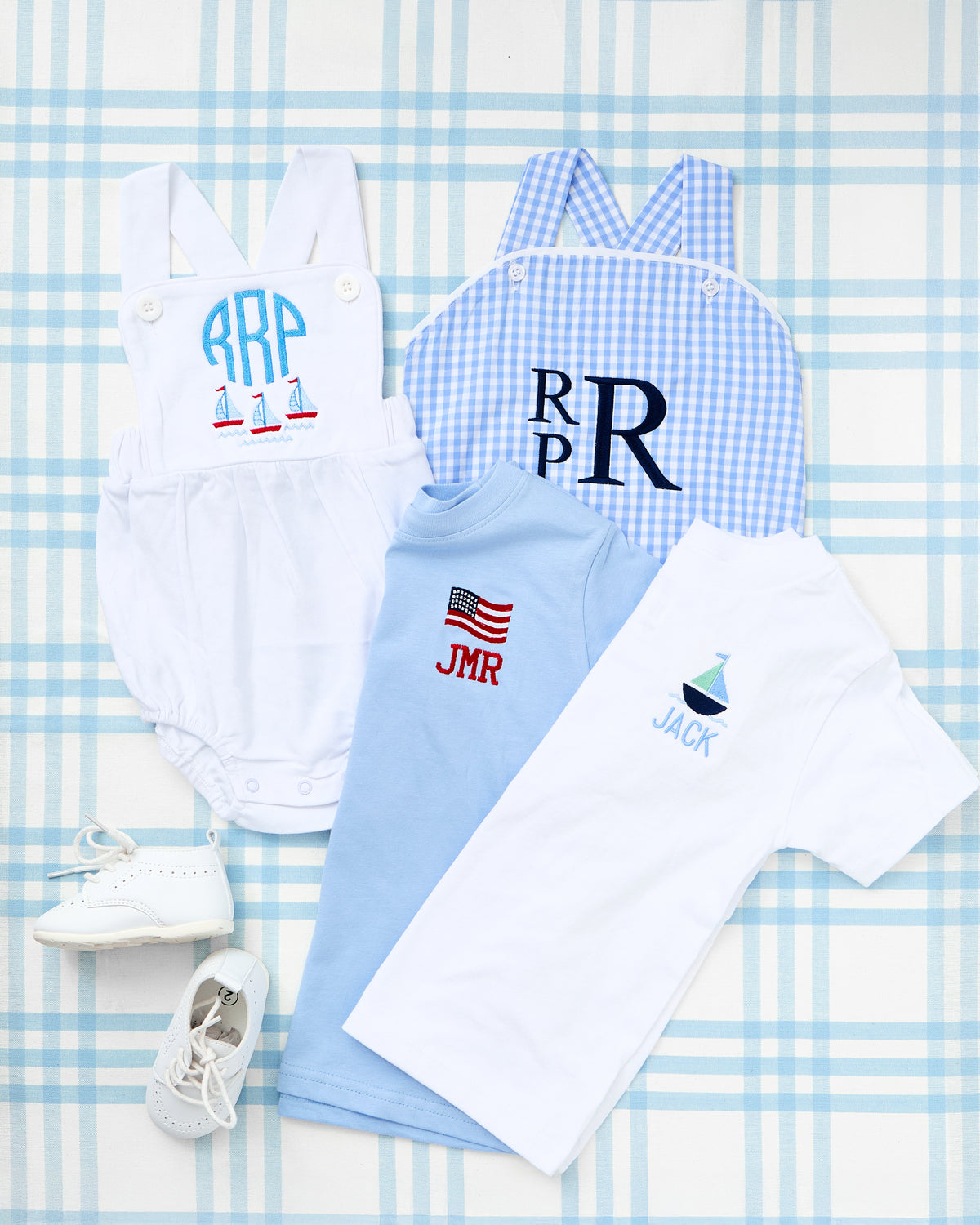Flat lay featuring personalized toddler outfits including two Solid Short Sleeve T-Shirts with Motif—one in light blue with an embroidered American flag and initials “JMR,” and one in white with a sailboat and name “JACK.” Also shown are a white cotton sunsuit with red sailboats and monogram “RRP,” a blue gingham sunsuit with bold initials “RPR,” and a pair of white baby shoes, all styled on a blue plaid background for a crisp, coastal Americana look.