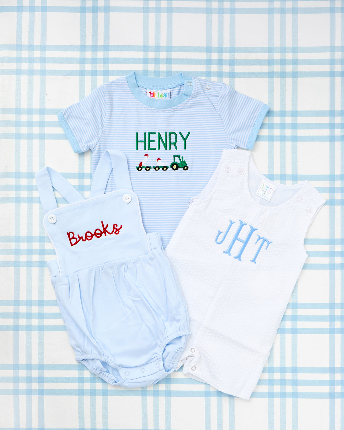 Flat lay image of three adorable baby outfits displayed on a blue gingham background, with a focus on the Cotton Sunsuit in the center. This light blue sunsuit features criss-cross straps and personalized red embroidery with the name “Brooks,” combining comfort with classic Southern charm. The set also includes a blue striped short sleeve bubble with a green tractor and the name “Henry,” and a white Seersucker Jon Jon with light blue monogram “JHT.” Perfect for warm weather play or keepsake moments.