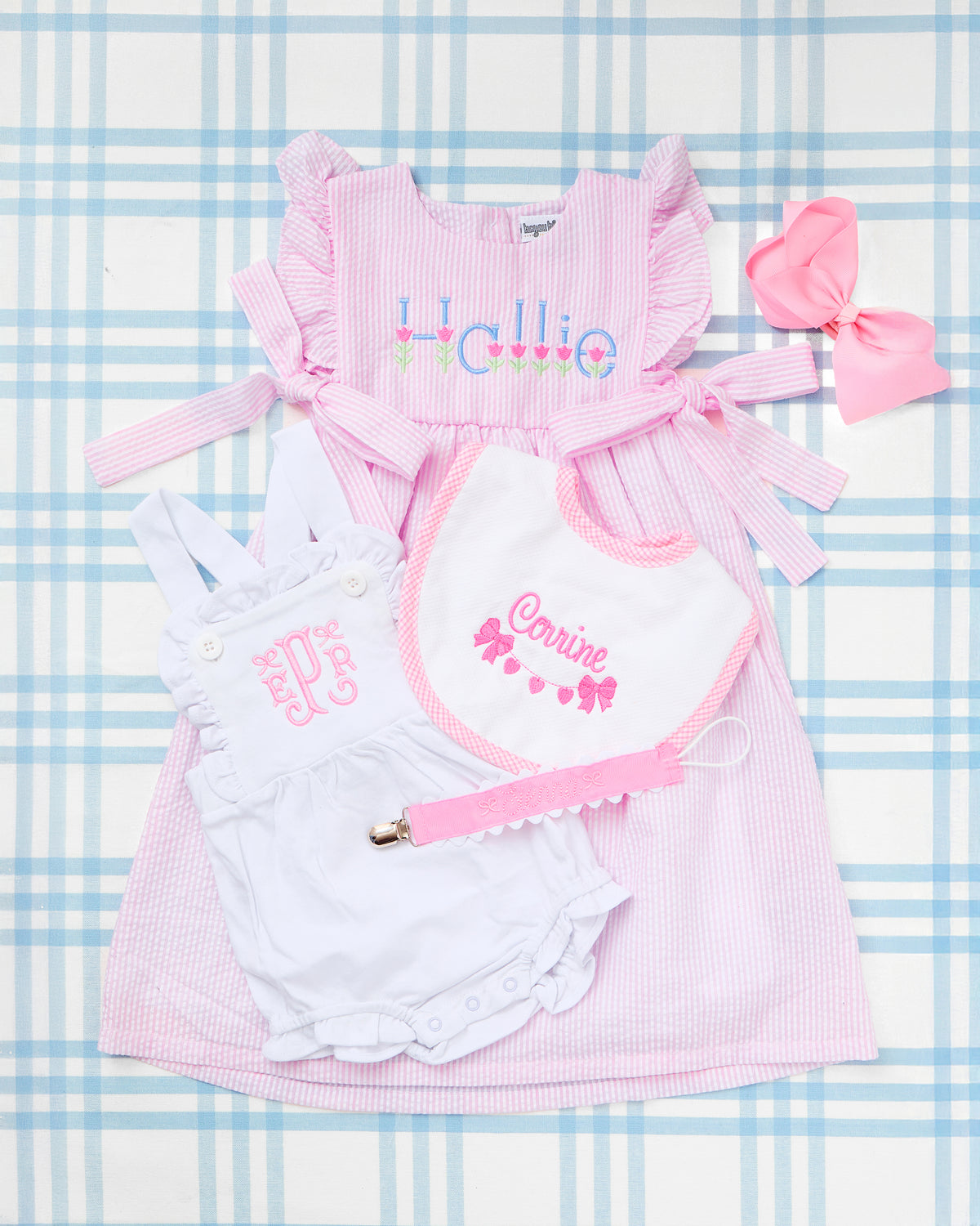 Flat lay of charming baby girl essentials displayed on a blue gingham background, featuring the standout Seersucker Bow Dress in pink with ruffle sleeves, side bows, and an embroidered tulip motif with the name “Hallie.” Paired with a white Cotton Sunsuit showcasing elegant pink monogramming, a coordinating embroidered bib with the name “Corinne” and bow motif, a pink pacifier clip, and a matching pink hair bow—this personalized set is perfect for springtime gifting or stylish everyday wear.