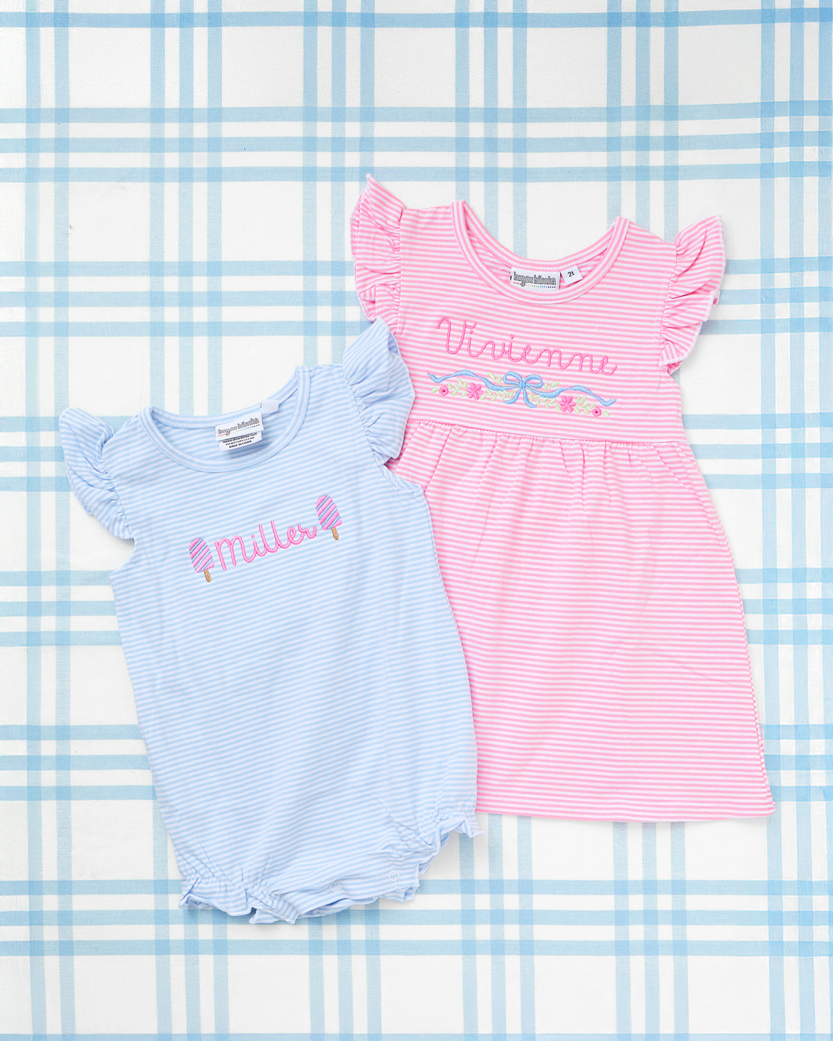 Personalized girls’ outfits from Rainey Laine featuring a light blue striped onesie with ruffle sleeves, pink popsicle motifs, and the name ‘Miller’ embroidered in pink, paired with a pink striped flutter-sleeve dress with floral bow motif and the name ‘Vivienne,’ displayed on a soft blue plaid background.