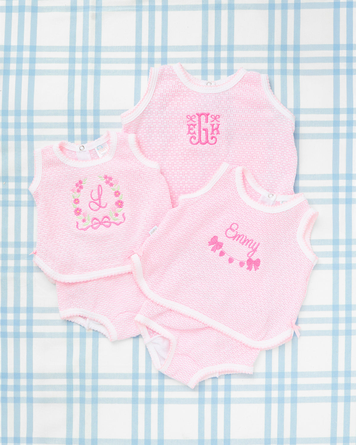 Flat lay of three Sleeveless Knit Diaper Cover Sets with Motif in soft pink, displayed on a blue plaid background. Each set features cozy textured knit fabric with white trim and custom embroidery: a floral wreath motif with the initial “L,” a monogram “EJG,” and a delicate bow banner design with the name “Emmy.” A charming and personalized baby girl outfit perfect for springtime photos or everyday sweetness.