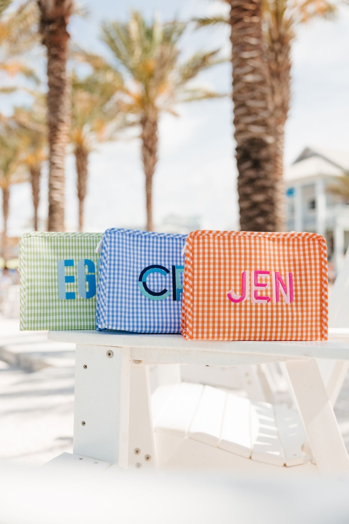 Three gingham pouches: one green with 'EG' monogram in teal, one blue with 'CM' monogram in navy, and one orange with 'JEN' monogram in pink.