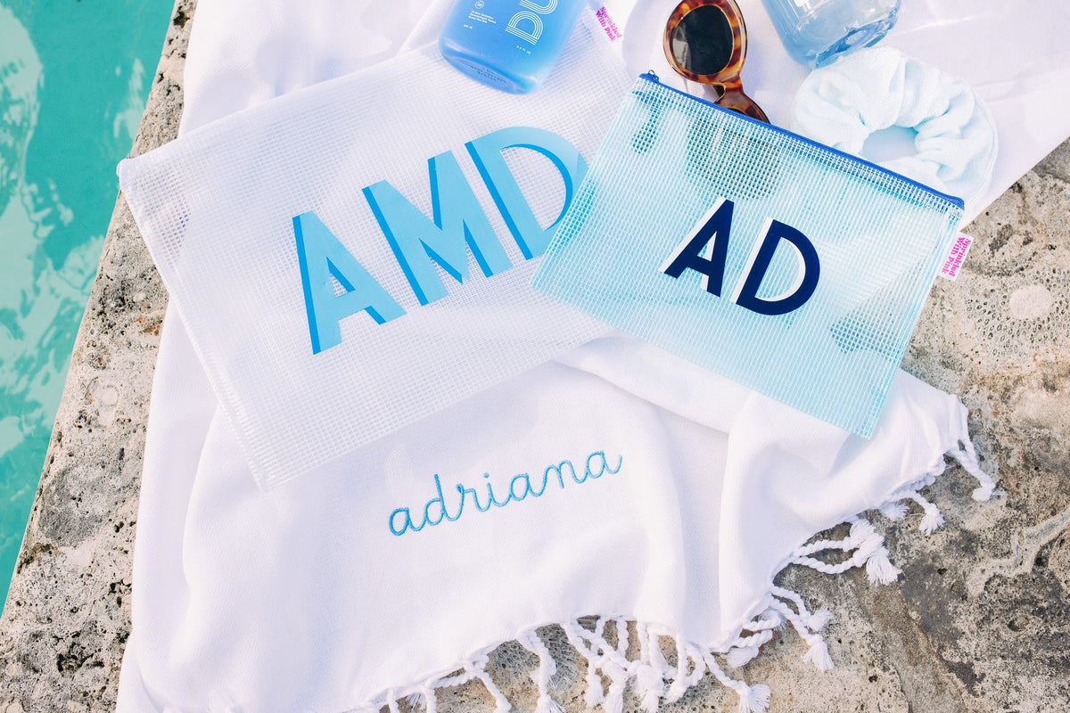 Flatlay of two Shadow Monogram Pool Bags beside a pool, personalized with initials “AMD” and “AD” in bold, vibrant shades of blue. The mesh-style pouches are ideal for beach or swim essentials and are shown alongside a monogrammed towel embroidered with the name “adriana,” sunglasses, scrunchie, and blue sunscreen bottle—all styled on a textured white backdrop for a clean, summery vibe.